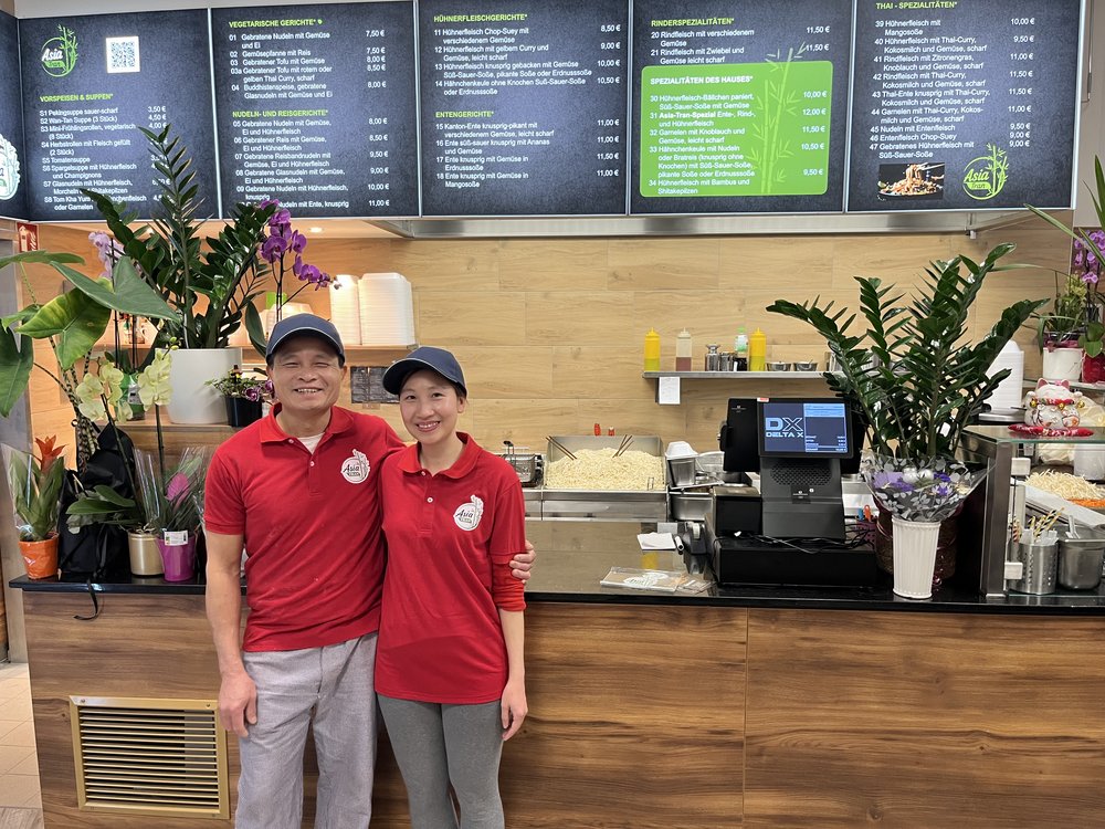 Zwei Personen in roten Shirts vor einer asiatischen Imbiss-Theke mit Speisekarte, Pflanzen und Kasse.